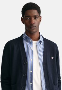 Dunkelblauer Cardigan mit Knöpfen über einem blauen und weißen gestreiften Hemd. Der Cardigan hat Taschen und ein Logo auf der Brust.