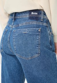 Jeans in denim blu con vita alta, design classico a cinque tasche e un'etichetta in tessuto scuro con la scritta "Brax" sulla parte posteriore della cintura.