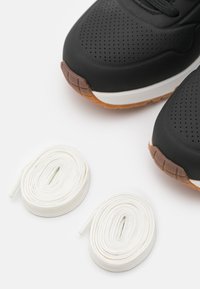 Black sneaker with perforated upper, white midsole, and brown rubber outsole. Two white laces positioned below the shoe.