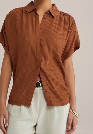 Person iført en tekstureret brun kortærmet knapoplukkelig skjorte, hvide bukser med sort bælte og et guldarmbånd på højre håndled.