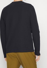 Personne portant une chemise noire à manches longues et un pantalon jaune moutarde, vue de dos, debout contre un fond blanc uni.