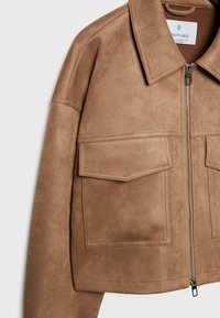 Veste en daim brun clair avec de grandes poches avant à rabat, fermeture éclair argentée et large col, présentée sur un fond clair.