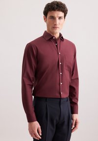 Bordeaux langermige shirt met een button-down kraag, voorzak en witte knopen, gecombineerd met donkere op maat gemaakte broeken. Zachte textuur.