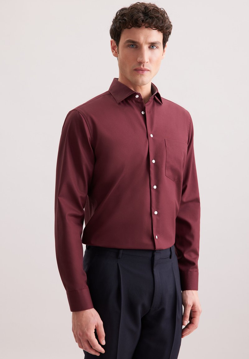 Bordeaux langermige shirt met een button-down kraag, voorzak en witte knopen, gecombineerd met donkere op maat gemaakte broeken. Zachte textuur.