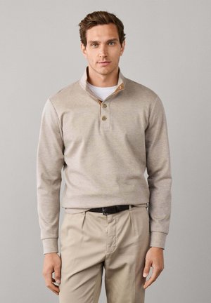 Man wearing beige long-sleeve pullover with buttons over white undershirt and light beige pants against plain gray background.