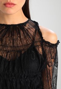 Indumento in pizzo nero con colletto alto, maniche lunghe trasparenti e spalle scoperte, caratterizzato da motivi intricati e una texture fluida.