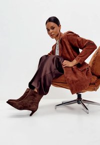 Barna velúr dzseki és sötétbarna szatén nadrág; a modell hegyes orrú, kúp sarkú bokacsizmát visel, modern székben ülve.