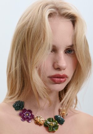 Collana colorata a forma di fiore, realizzata in plastica e metallo, con accenti in viola, verde e oro, indossata al collo di un modello.