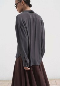 Blusa a maniche lunghe grigia con colletto e polsini abbottonati; abbinata a una gonna marrone fluida. Tessuto liscio con una leggera lucentezza.