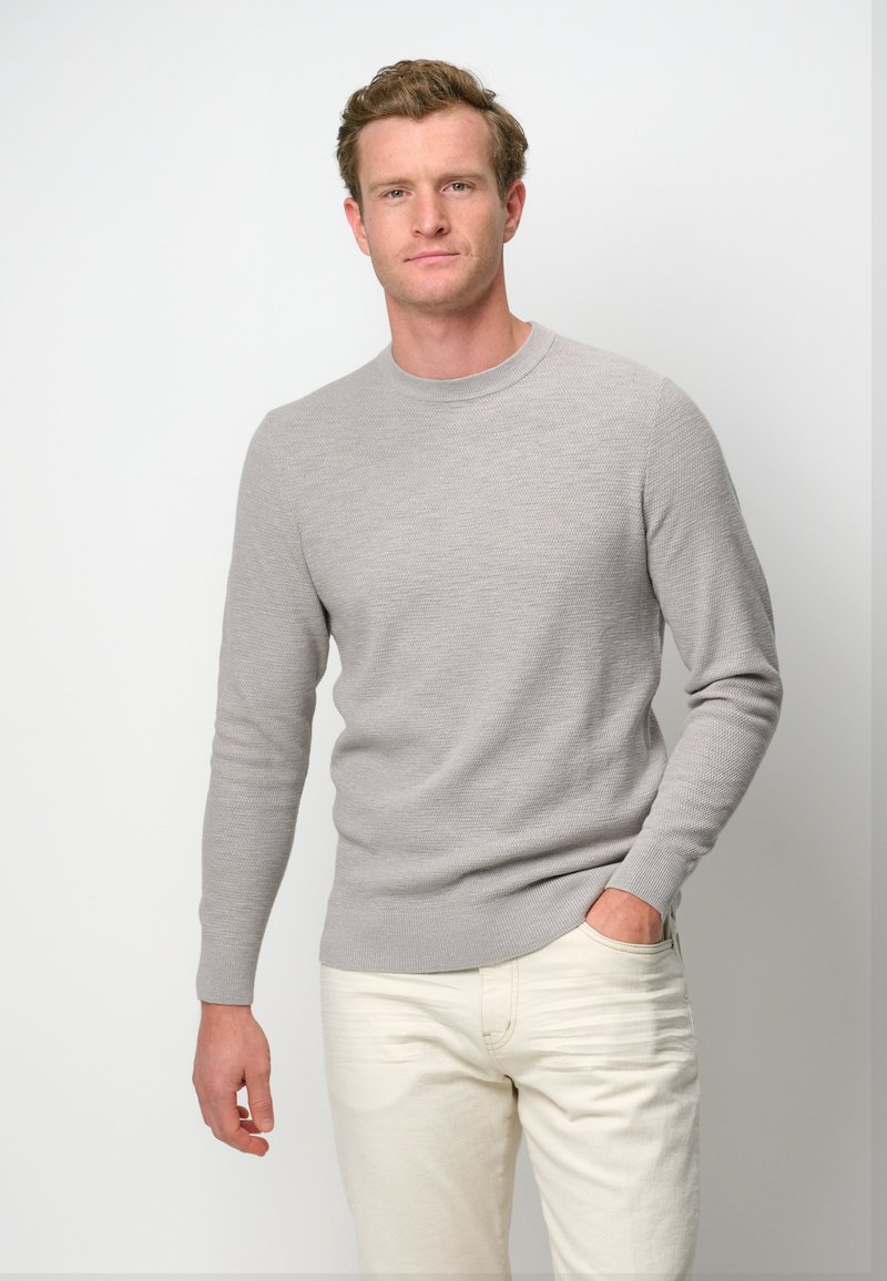 Junger Mann mit kurzen lockigen Haaren trägt einen hellgrauen Strickpullover und elfenbeinfarbene Hose, eine Hand in der Tasche.