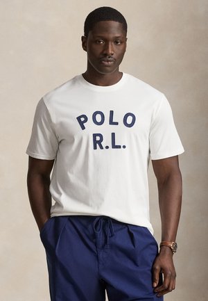 Férfi fehér rövid ujjú pólót visel, amelyen "POLO R.L." felirat látható tengerészkék színben, valamint tengerészkék megkötős nadrágot, kezét zsebre téve áll.