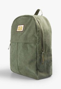 Sac à dos en toile vert olive avec poche avant, poignée supérieure, fermeture éclair latérale et un écusson rectangulaire avec texte jaune sur la face avant.