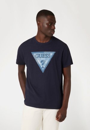 Camiseta de algodón azul marino con un gráfico triangular de color azul claro que presenta el logo "GUESS" y un detallado de costura. Mangas cortas, ajuste casual.