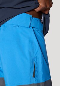 Blå shorts lavet af letvægtsstof med en tætsiddende talje. Har sidelommer med sort lynlås og en kontrasterende grå kant.