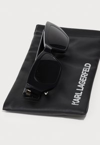 Svart, kantete solbriller laget av plast, hvilende på en svart lærpung med innpreget "KARL LAGERFELD" i hvitt.