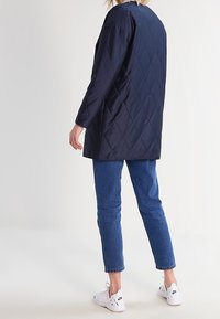 Osoba oblečená v námořnické prošívané bundě, modrých džínách a bílých teniskách Nike, stojí s zády otočenými kameře na jednolitém pozadí.