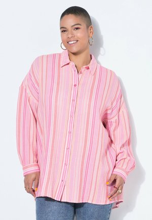 Lächende Person mit kurzen Haaren, die große Creolen trägt und ein lockeres, pink-gestreiftes Hemd mit Knöpfen sowie blaue Jeans anhat.