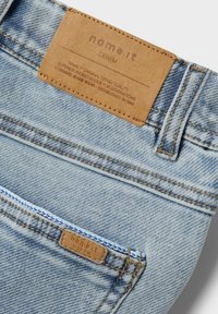 Svijetloplave traperice s kožnom oznakom koja glasi "name it DENIM." Sadrže vidljive šavove i detalje na džepovima, ističući teksturirani materijal.