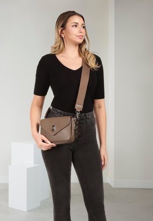 Femme portant un haut et un pantalon noirs, tenant un sac bandoulière taupe avec une large sangle, debout dans une pièce aux tons neutres avec des blocs d'exposition blancs.