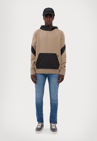 Felpa beige con accenti neri e una tasca frontale, abbinata a jeans blu e sneakers grigie. Il modello indossa un cappellino nero. Design semplice.