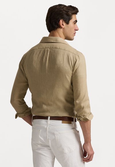 Beżowa lniana koszula z podwiniętymi rękawami, detal z plisą z tyłu, w połączeniu z białymi spodniami i brązowym paskiem, eksponująca teksturowany materiał.