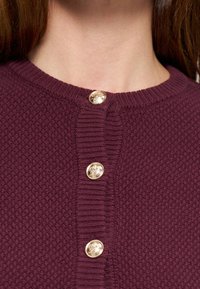 Gros plan d’un cardigan en maille texturée bordeaux avec trois boutons dorés embossés le long de la patte de boutonnage près de l’encolure.