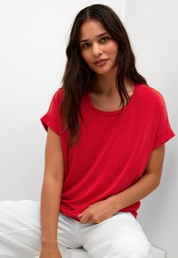 Maglietta rossa a maniche corte con scollo rotondo, texture morbida e design drappeggiato. Abbinata a pantaloni bianchi larghi, seduta su uno sfondo chiaro.