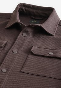 Veste en tissu brun foncé texturé avec col, boutons pression sur le devant et poche à rabat sur la poitrine.