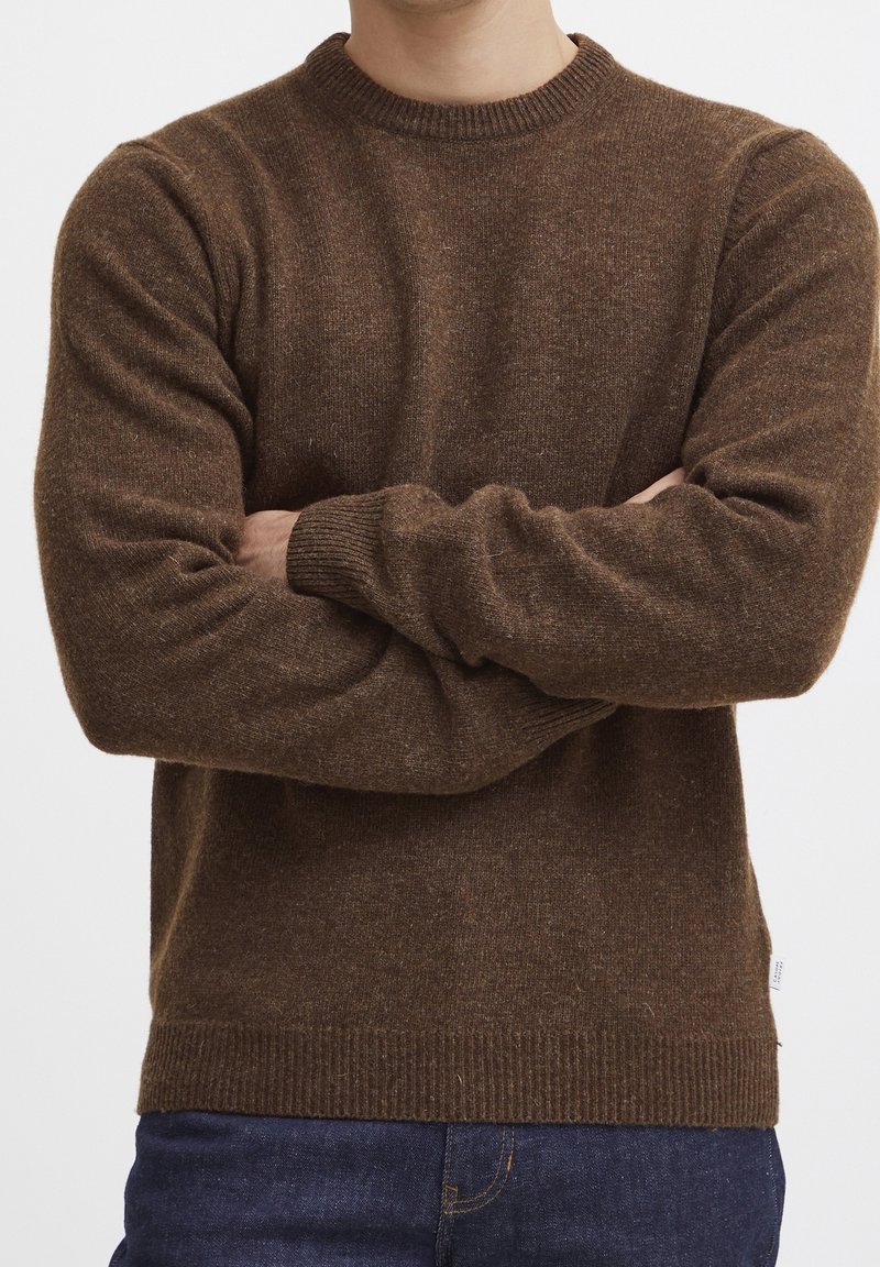 Homme portant un pull en maille marron à col rond, bras croisés, associé à un jean bleu foncé, vu du cou jusqu'aux hanches sur fond blanc.