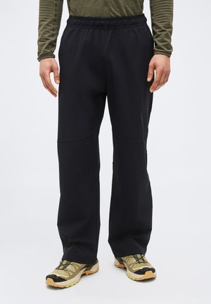 Férfi fekete bővebb szabású nadrágban, olívazöld hosszú ujjú felsőben és bézs túracipőben, egyszínű háttér előtt áll.