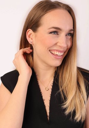 Gouden hartvormige ketting om de nek van een vrouw met lang haar, die een zwart topje met rafels draagt, lachend in een heldere omgeving. Zilveren oorbellen zichtbaar.