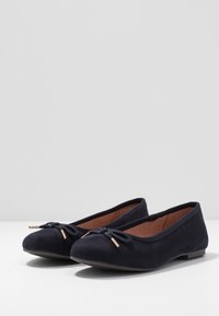 Ein Paar schwarze Wildleder-Balletschuhe mit runden Zehen und einer dekorativen geflochtenen Schleife mit metallischen Spitzen auf einer weißen Oberfläche.