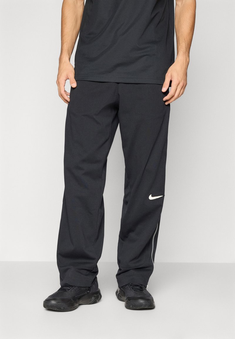 Melnas sporta bikses ar brīvu piegriezumu, kurām pa kreisi pie kājas ir balts Nike logotips un apakšā ir rāvējslēdzēji, lai varētu pielāgot atveres.