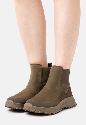 Bottes chevilles en suede vert avec panneaux latéraux élastiques, semelle en caoutchouc texturée et tirant à l'arrière pour un enfilage facile.