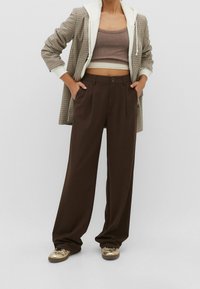 Brązowe spodnie o szerokich nogawkach z dopasowanym krojem, z zapięciem na guzik i dwoma bocznymi kieszeniami, w zestawieniu z długim marynarskim płaszczem w kratę oraz beżową crop top.