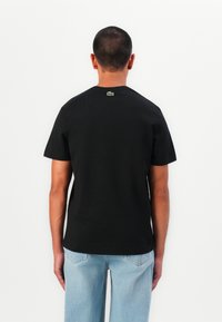 Sort bomulds t-shirt med korte ærmer, rund halsudskæring og et lille grønt logo på den øverste ryg. Kombineret med lys blå vide ben jeans.