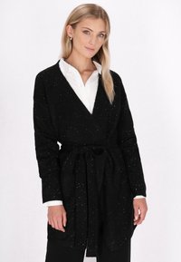 Zwarte cardigan met een strik in de taille, lange mouwen en een wit gespikkelde textuur. Draagt over een witte overhemd met kraag, wat een casual gelaagde look toont.