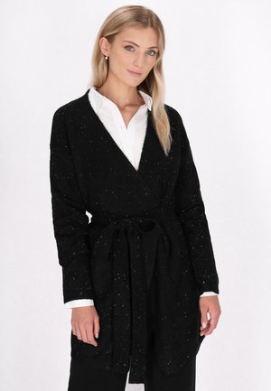 Schwarzer Cardigan mit Bindegürtel, langen Ärmeln und weiß gesprenkelter Textur. Über einem weißen Hemd mit Kragen getragen, das einen lässigen Lagenlook zeigt.