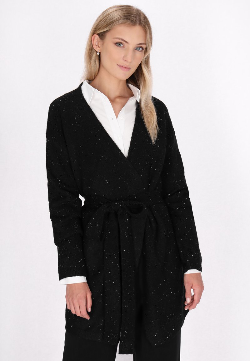 Zwarte cardigan met een strik in de taille, lange mouwen en een wit gespikkelde textuur. Draagt over een witte overhemd met kraag, wat een casual gelaagde look toont.