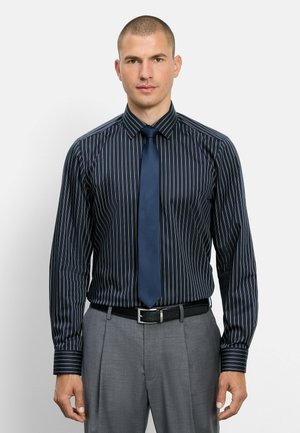 Uomo che indossa una camicia nera a righe sottili con una cravatta blu navy e pantaloni grigi, in piedi davanti a uno sfondo chiaro uniforme.