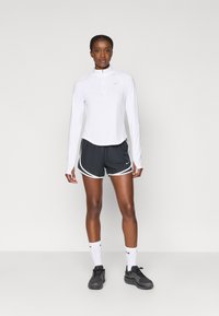 Λευκό αθλητικό μπλουζάκι με μακριά μανίκια και φερμουάρ στο κολάρο, συνδυασμένο με μαύρα σορτς με λευκές ρίγες πλευρικά και μαύρα αθλητικά παπούτσια με γκρίζες λεπτομέρειες.