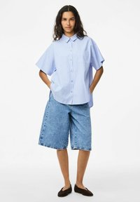 Gaiši zila svītru īsa piedurkņu krekls ar apkakli un kabatu, apvienots ar vaļīgām džinsa šortiem. Modelis stāv ar rokām kabatās.