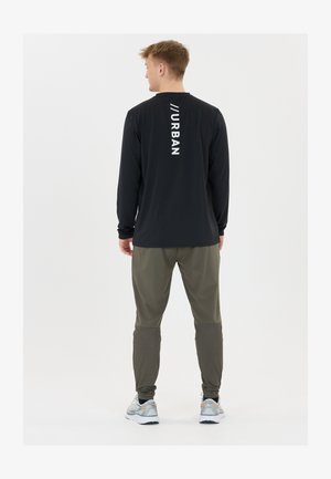 Mies, который стоит спиной, на нем черная рубашка с длинным рукавом с надписью "URBAN" на спине, оливковые джоггеры и белые кроссовки для бега.