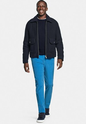 Glunderende man die wandelt, gekleed in een marineblauwe jas met ritssluiting, een zwart shirt, felblauwe broek en zwarte sneakers met witte zolen tegen een effen achtergrond.