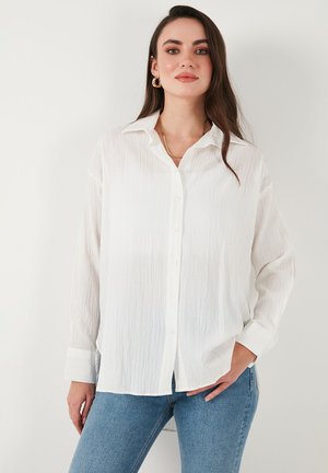 White button-up shirt with a slightly wrinkled texture, long sleeves, and a collar, paired with blue jeans.