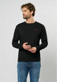 Schwarzes Langarmhemd in taillierter Form, mit einem kleinen roten Logo auf der oberen linken Seite, kombiniert mit hellblauen Jeans.