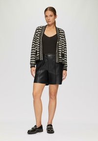 Zwart-wit geblokte cardigan met gouden knoppen, zwarte tanktop en zwarte leren shorts. Glanzende leren schoenen maken de outfit compleet.