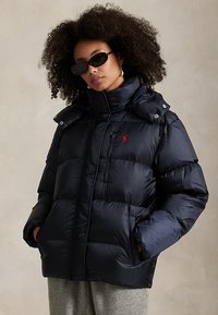 Frau mit lockigem Haar, die schwarze Sonnenbrillen und eine schwarze Daunenjacke mit rotem Logo trägt, Hände in den Taschen, vor einem schlichten Hintergrund stehend.