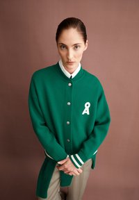 Cardigan vert à boutons avec un col rond, des poignets rayés blancs et une lettre "A" brodée en blanc sur la poitrine.