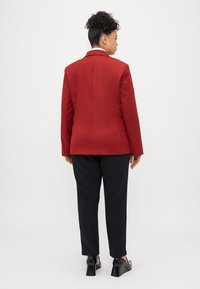 Raudonas švarkas su struktūrizuotu dizainu, vienu vents užpakalyje, derinamas su juodomis kelnėmis ir aukštakulniais batais. Lygus tekstūros paviršius, minimali detalizacija.